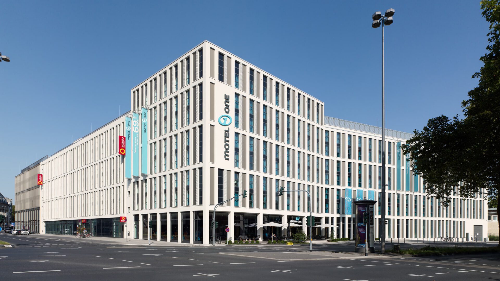 Motel One bei Tag, die Gebäudeecke mit den Bäumen der Straße