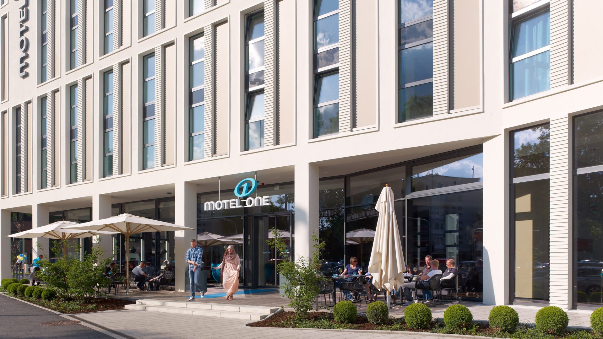 Terrasse vor dem Motel One mit Gästen unter Sonnenschirmen